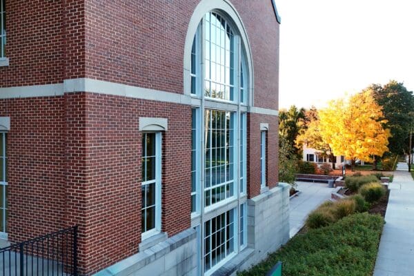 Close-up exterior of a Nichols College brick building with a large arched window, with a vibrant yellow fall foliage tree visible along the campus sidewalk