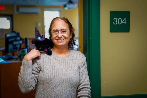 Professor Emily Thomas leans in the doorway of Room 304 in Academy Hall at Nichols. On her shoulder rests her black cat plush toy Geillis Duncan, which she named after the young maidservant in 16th century Scotland accused of witchcraft