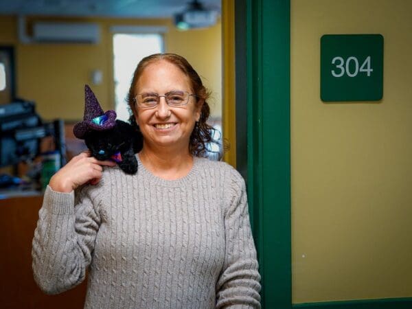 Professor Emily Thomas leans in the doorway of Room 304 in Academy Hall at Nichols. On her shoulder rests her black cat plush toy Geillis Duncan, which she named after the young maidservant in 16th century Scotland accused of witchcraft