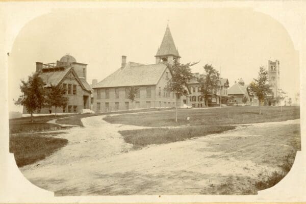 A historic black-and-white photograph of early campus buildings at Nichols College, showing brick structures and open grounds from the late 19th century.