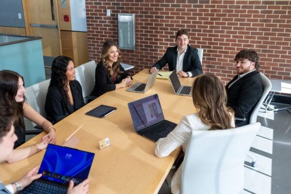 Nichols College students in professional attire collaborate around a conference table with laptops during a Bison Consulting Group meeting.
