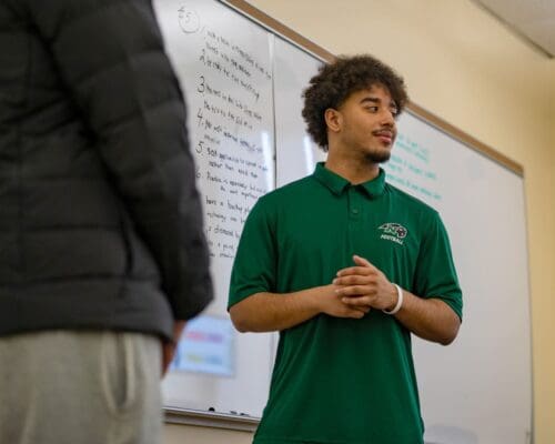 A Nichols College student in a green football polo presents in front of a whiteboard during a class at Nichols College.