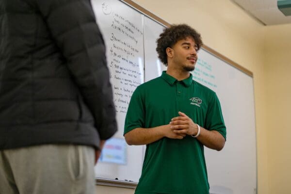 A Nichols College student in a green football polo presents in front of a whiteboard during a class at Nichols College.