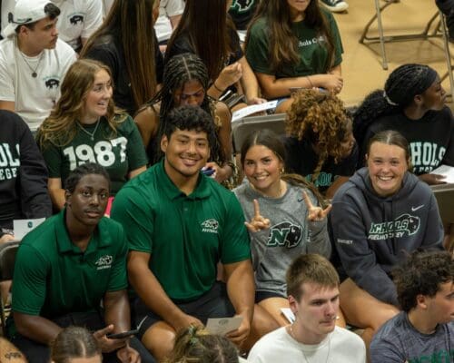 Nichols College students in school spirit apparel smile and cheer during a freshman orientation assembly on campus.