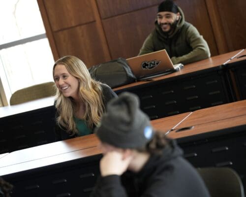 Students laugh and engage in discussion during a class in a wood-paneled lecture room on the Nichols College campus.