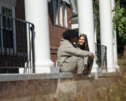 Two students sit and chat on the steps of a white-columned brick building on the Nichols College campus on a sunny fall day.