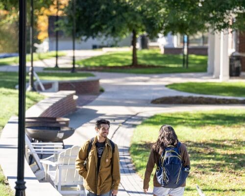 Two students with backpacks walk and chat along a tree-lined campus path at Nichols College on a fall afternoon.