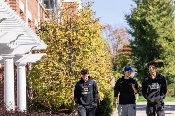 Three students with backpacks walk and smile along a campus path at Nichols College on a sunny fall day.