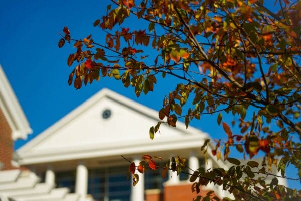 Autumn leaves frame the white pediment roofline of a brick building on the Nichols College campus against a bright blue sky.