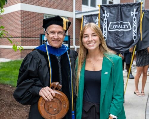 A faculty member in academic regalia holding the Nichols College mace poses with a student in a green blazer during the commencement ceremony.