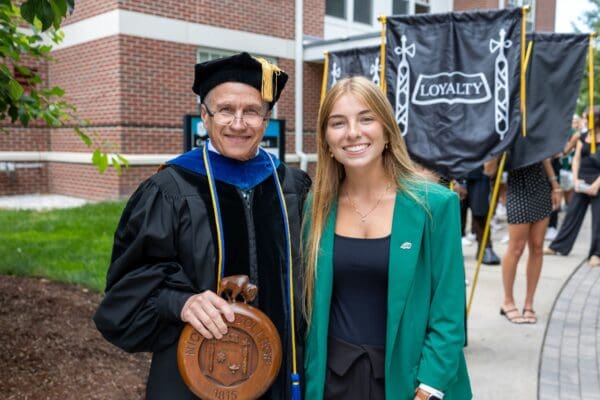 A faculty member in academic regalia holding the Nichols College mace poses with a student in a green blazer during the commencement ceremony.