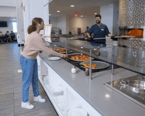 A Nichols College student selects pizza at the dining hall pizza station while a staff member serves fresh slices from a wood-fired oven.
