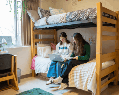 Two Nichols College students sit on a bed in a residence hall room, working together on a laptop.