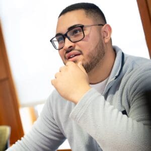 A student sitting at a table on Nichols' campus, wearing glasses and a gray fleece, listening attentively during a class or advising session.