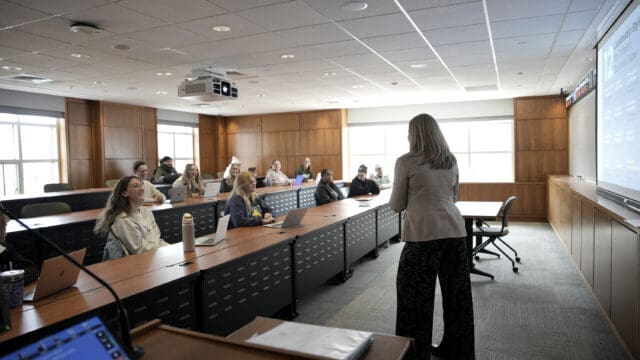 A professor leads a lecture for a class of engaged Nichols College students in a wood-paneled classroom, with a presentation projected on screen.