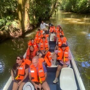 Nichols College students and faculty wearing orange life jackets ride a boat through a lush jungle waterway during a study abroad trip to Costa Rica.