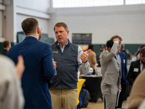 A man wearing a gray "Noble" branded vest and a plaid shirt gestures with a thumbs-up while speaking to a student in a blue suit during a crowded campus networking event.