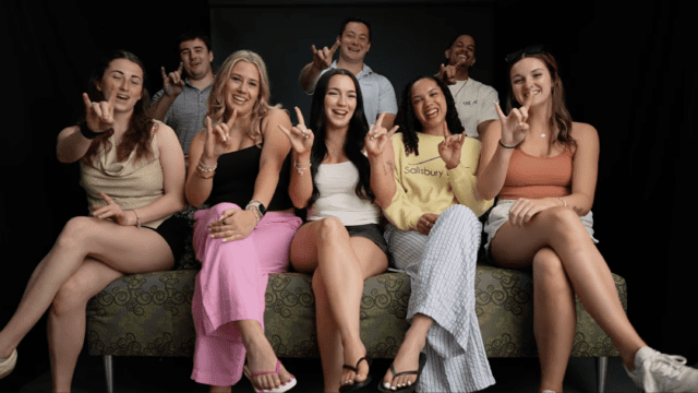Eight Nichols College seniors sit together on a couch smiling and making the hand sign "horns" to imitate the Bison, the Nichols mascot