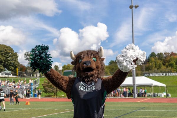 The Nichols College mascot, a brown bison wearing a black "Nichols College" tank top, stands on an athletic field holding a green pom-pom in one hand and a white pom-pom in the other.