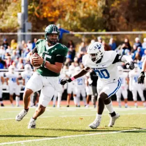 A dynamic action shot of a football game where a quarterback in a green Nichols College jersey runs with the ball while being pursued by a defender in a white jersey.