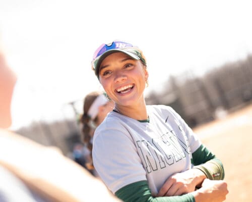 A smiling Nichols College softball player in uniform and cap laughs during an outdoor practice or game
