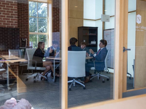 Four Nichols College students in business attire collaborate around a conference table in a glass-walled meeting room