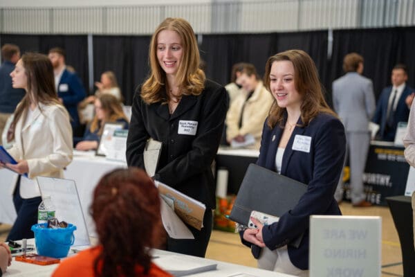 Two Nichols College students in professional attire smile while speaking with a recruiter at a career and internship fair booth