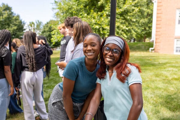 Two students smiling together on Nichols College campus lawn with other students gathered in the background.