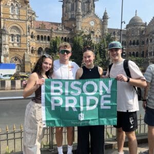 Four Nichols College students hold a green Bison Pride flag in front of Chhatrapati Shivaji Maharaj Terminus in Mumbai, India