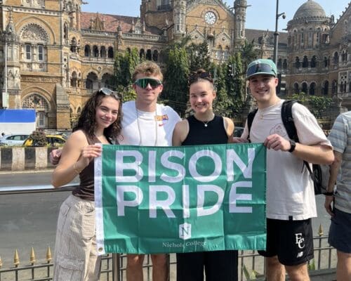 Four Nichols College students hold a green Bison Pride flag in front of Chhatrapati Shivaji Maharaj Terminus in Mumbai, India