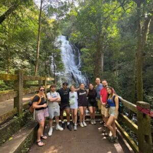 Nichols College students and faculty pose together on a bridge in front of a tropical waterfall surrounded by lush forest