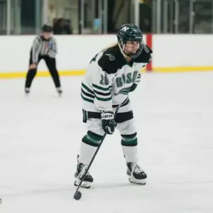 A Nichols College women's ice hockey player in a white, green, and black "Bison" jersey and full gear, poised with her stick on the ice during a game.
