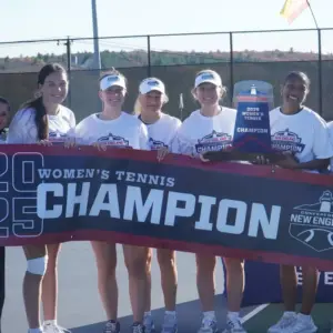 A group of Nichols College women's tennis players in white "Conference of New England Champion" shirts holding a large vinyl "2025 Women's Tennis Champion" banner and a trophy.